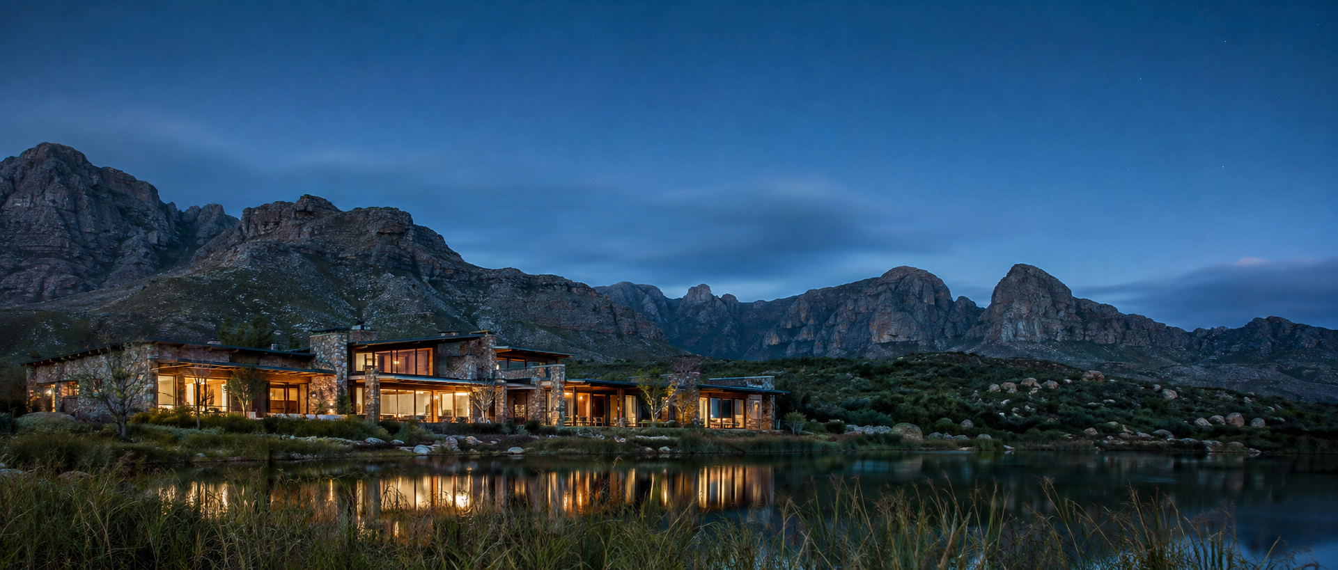 Dusk over still water below mountains, with a low lit building on the shore